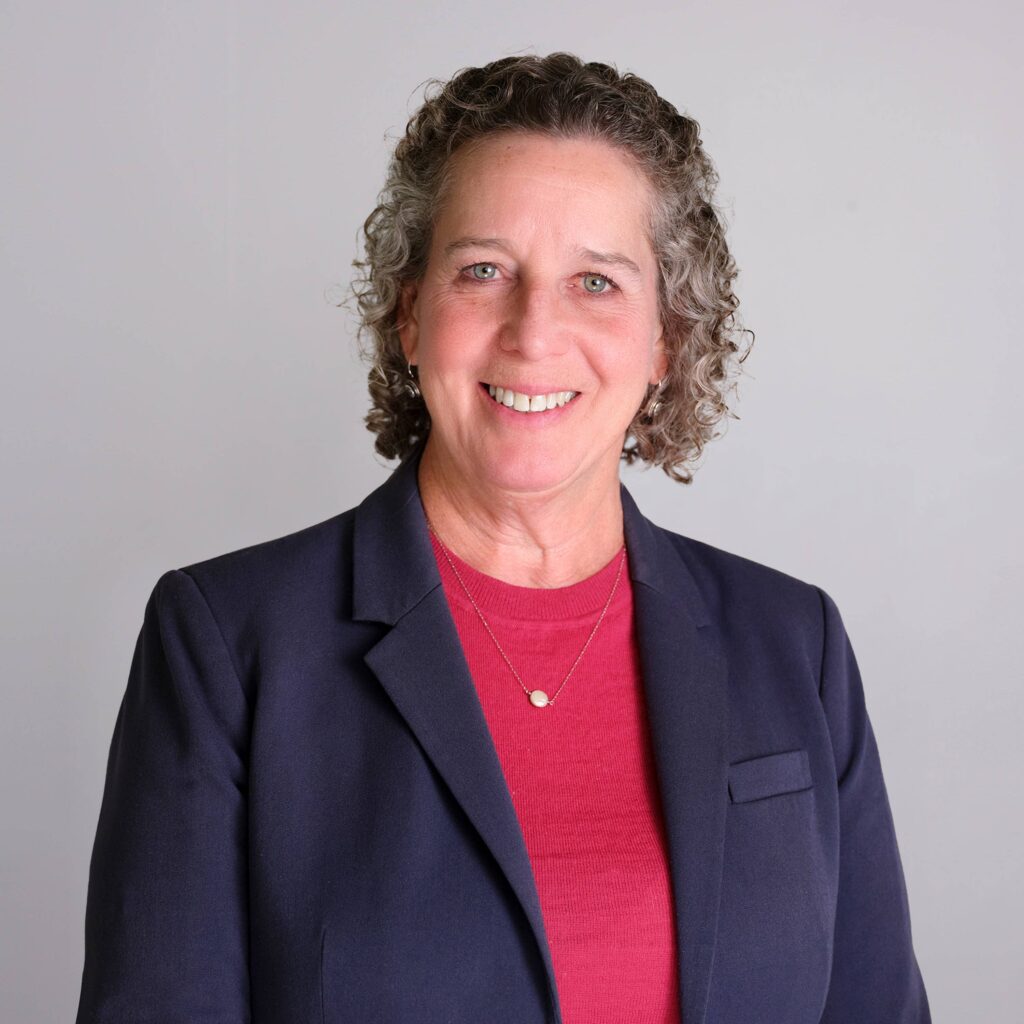 A person with curly gray hair smiles while wearing a navy blazer over a red shirt. The background is plain and light-colored.