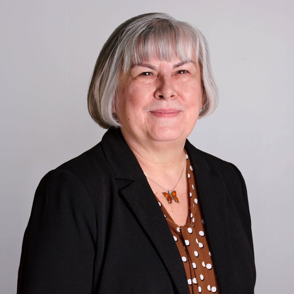 A woman with gray hair wears a black blazer over a brown polka dot blouse, accessorized with a butterfly necklace. She stands against a plain gray background, smiling gently at the camera.