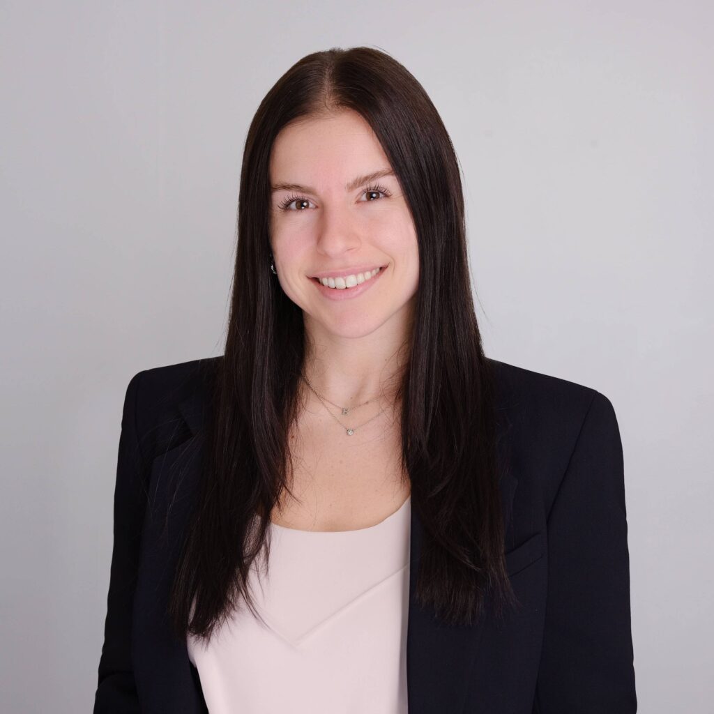 Brooklyn Seal with long, dark hair is smiling at the camera. She is wearing a white top and a dark blazer against a plain background.