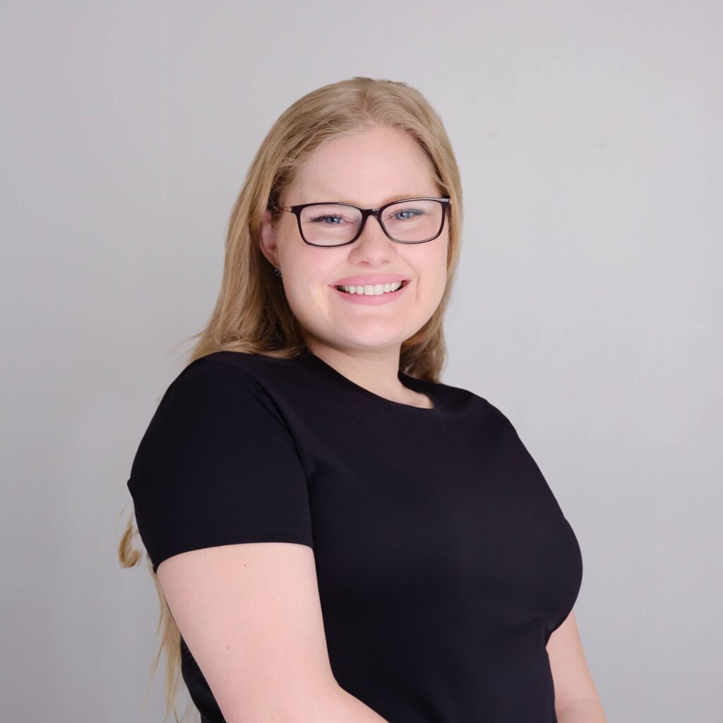 A person with long blonde hair and glasses smiles while wearing a black shirt. The background is plain gray.