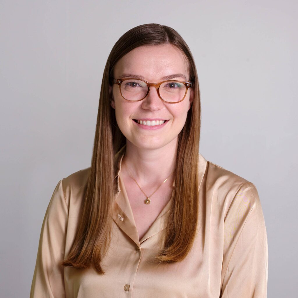 Hannah Leclair with long straight hair and glasses smiles at the camera, wearing a beige blouse. The background is plain light gray.