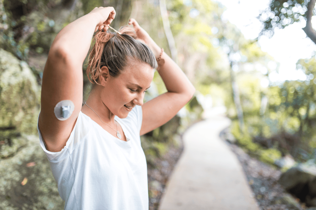 A woman stands on a path surrounded by greenery, smiling while tying her hair up. She wears a white shirt and has a medical device attached to her upper arm. The pathway curves into the distance through the trees.