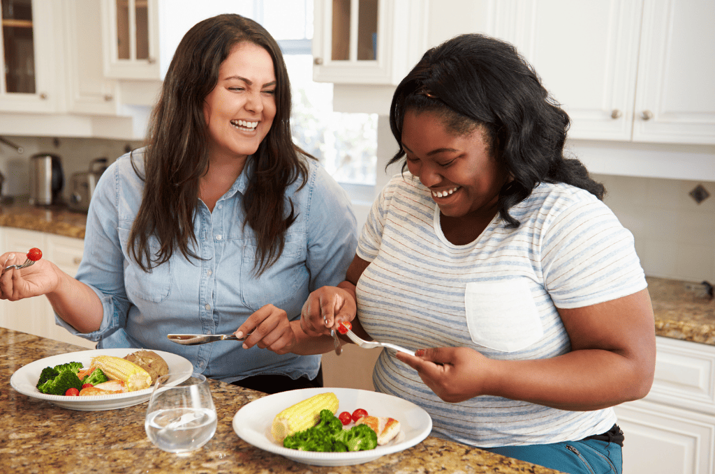 Two women sitting at a kitchen counter, enjoying a meal together. They are laughing and appear to be sharing a light moment. Plates in front of them contain salad, corn, and a potato. A glass of water is on the counter.