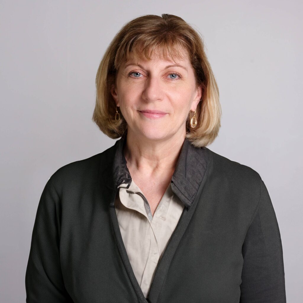 A woman with short blond hair is wearing a dark gray jacket over a beige collared shirt. She is standing against a plain, light gray background and looking at the camera with a slight smile.