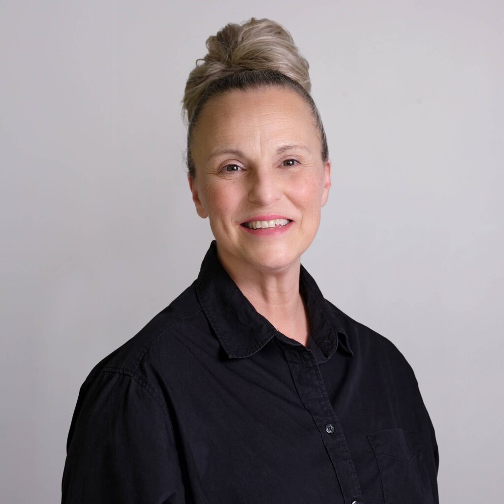 Jenny Schiffl with blonde hair in a bun smiles at the camera. She is wearing a black button-up shirt and stands against a plain light gray background.