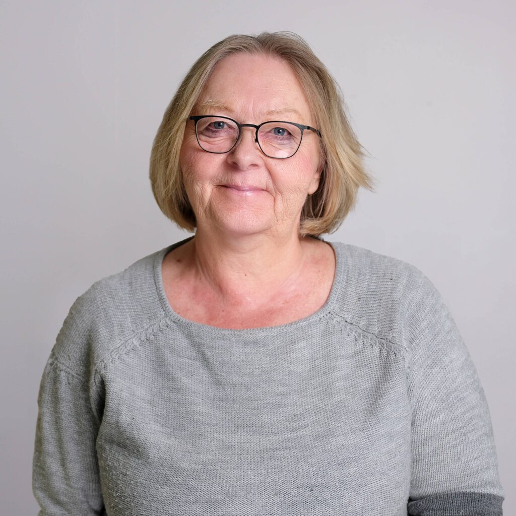 An older woman with short blonde hair and glasses smiles at the camera. She is wearing a light gray sweater, standing against a plain light gray background.