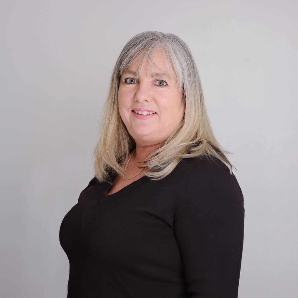 A woman with shoulder-length gray hair is smiling at the camera. She is wearing a black top and standing against a plain light gray background.