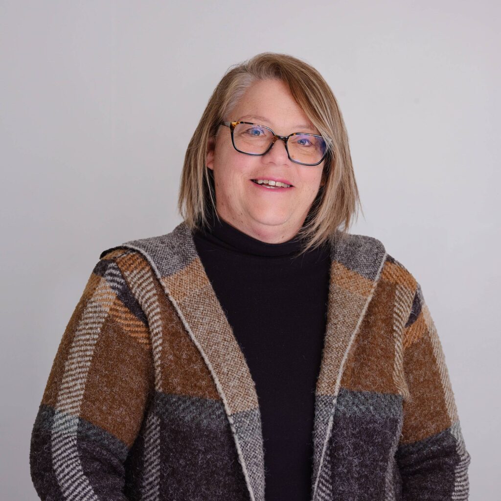 A person with shoulder-length hair and glasses is smiling. They are wearing a patterned brown and beige cardigan over a black turtleneck. The background is plain and light-colored.