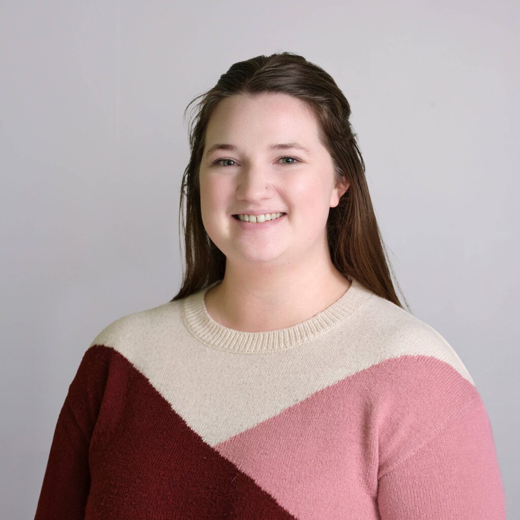 Portrait of a person smiling, wearing a sweater with color blocks of beige, maroon, and pink. They have long brown hair and are standing against a plain light gray background.