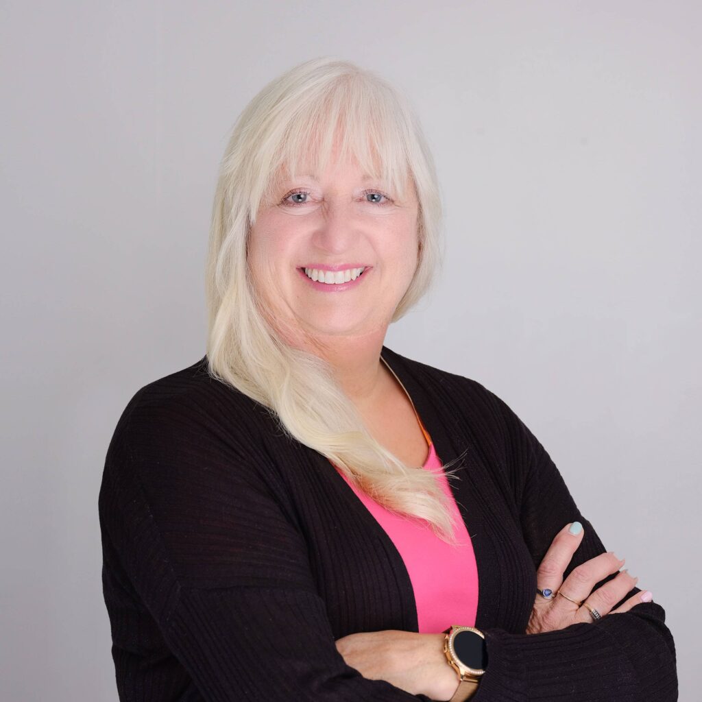 A person with long white hair is smiling, standing with arms crossed. They are wearing a black cardigan and a pink shirt, and have a smartwatch on their wrist. The background is light gray.