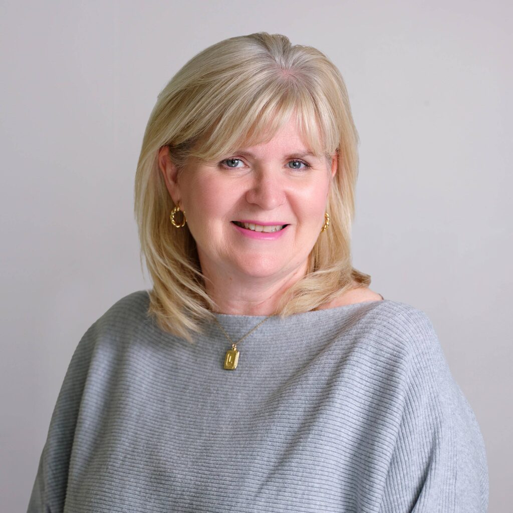 A woman with shoulder-length blonde hair smiles at the camera. She is wearing a light gray top, gold hoop earrings, and a gold necklace. The background is plain light gray.