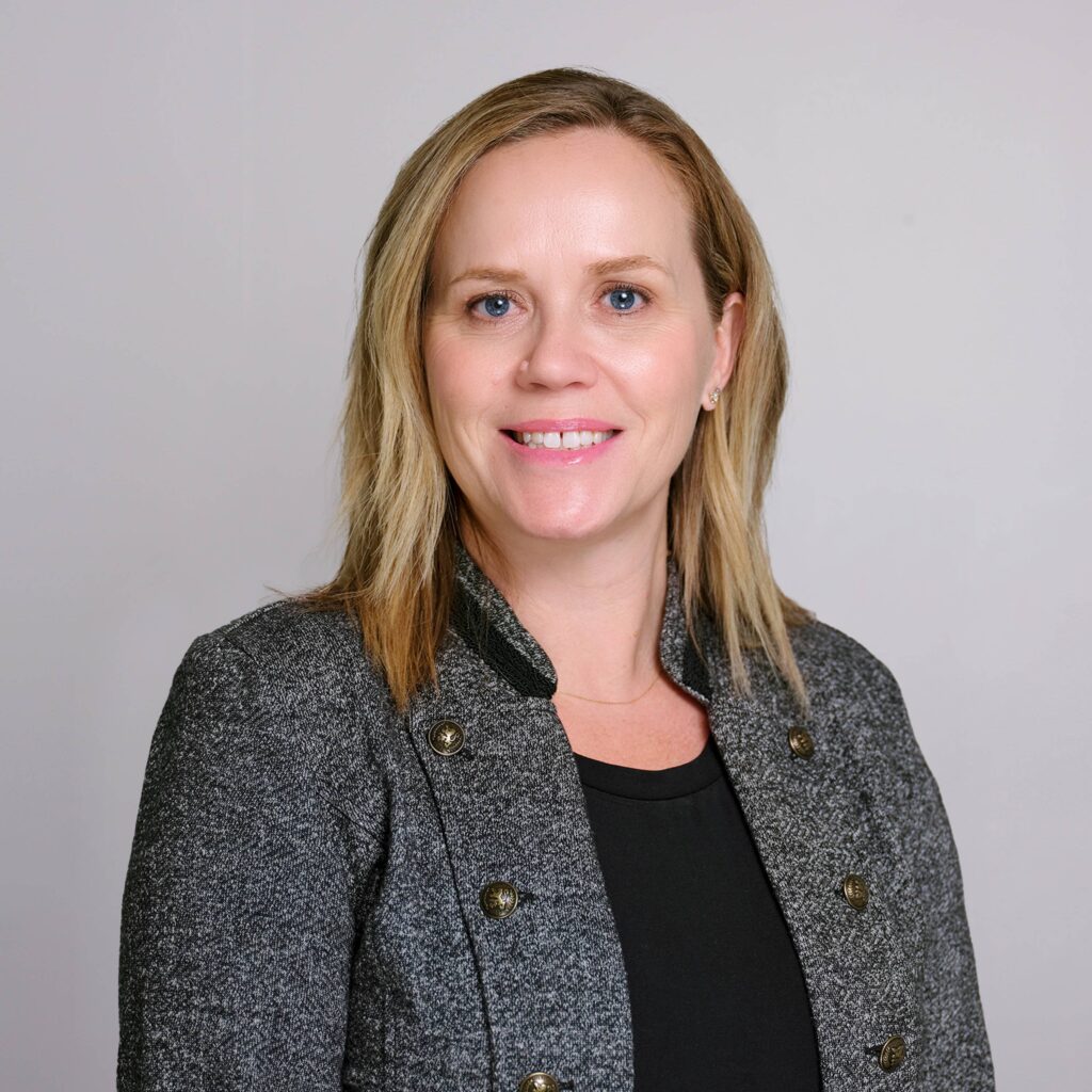 A woman with shoulder-length blonde hair smiles, wearing a grey blazer with black trim and a black top. The background is plain and light-colored.