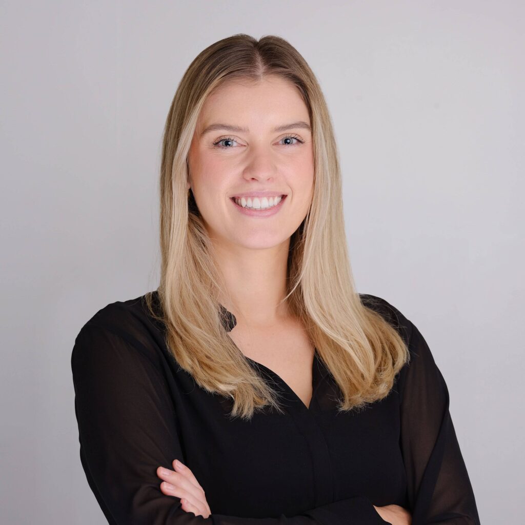 A woman with long blonde hair wearing a black blouse smiles confidently with her arms folded against a plain gray background.