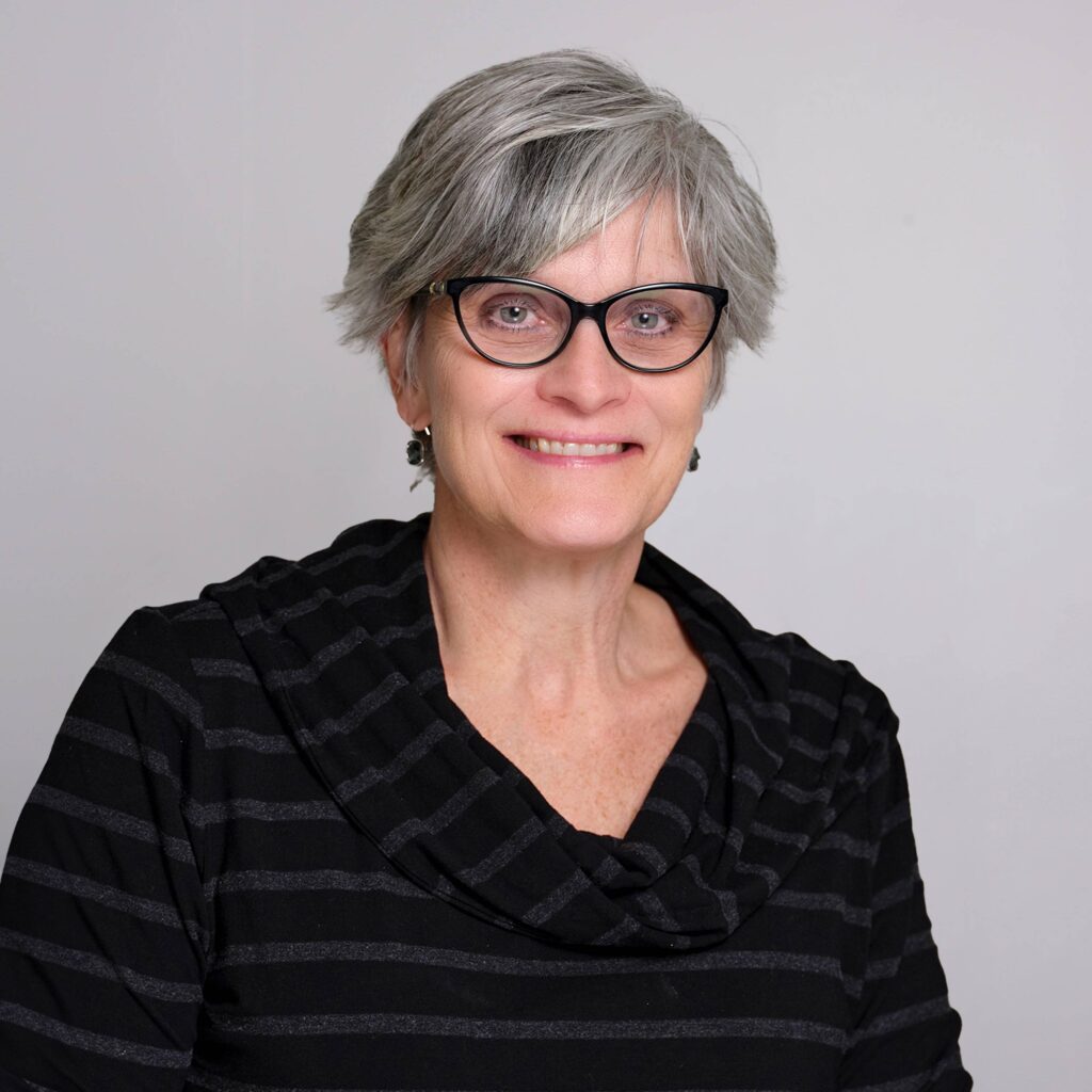 An older woman with short gray hair and black-framed glasses smiles at the camera. She is wearing a black and gray striped top against a plain gray background.