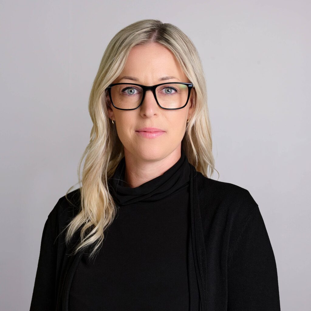 A person with long blonde hair and glasses is wearing a black top and cardigan. They are standing against a plain light gray background, looking directly at the camera with a neutral expression.