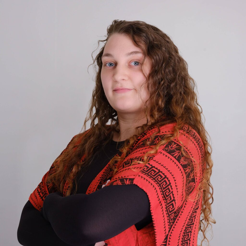 A person with long curly hair stands against a plain background, wearing a red patterned shawl over a black top, looking confidently at the camera with arms crossed.