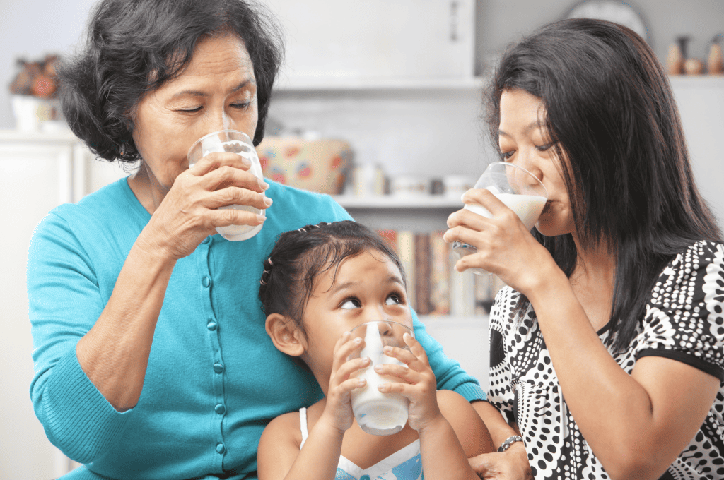 An older woman, a young woman, and a child sit together in a kitchen, each holding a glass of milk. The older woman wears a blue sweater, the younger woman a black-and-white patterned top, and the child has a white shirt. They are smiling and drinking.