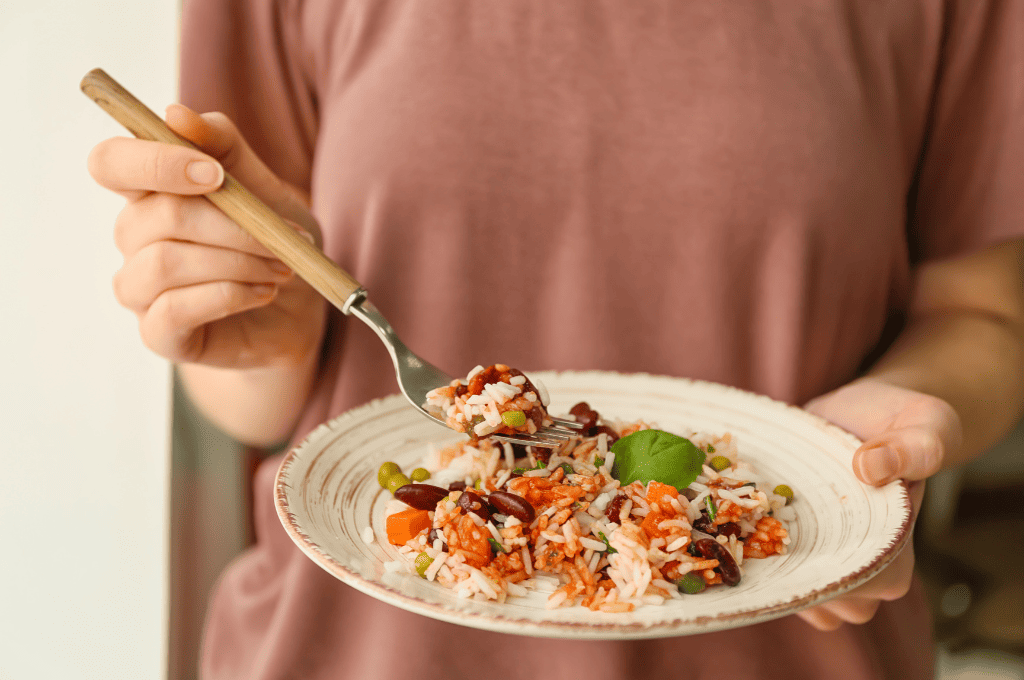 A person in a mauve shirt holds a plate of rice mixed with beans, peas, and carrots. A fork with the same food is in the other hand. A basil leaf garnishes the dish.