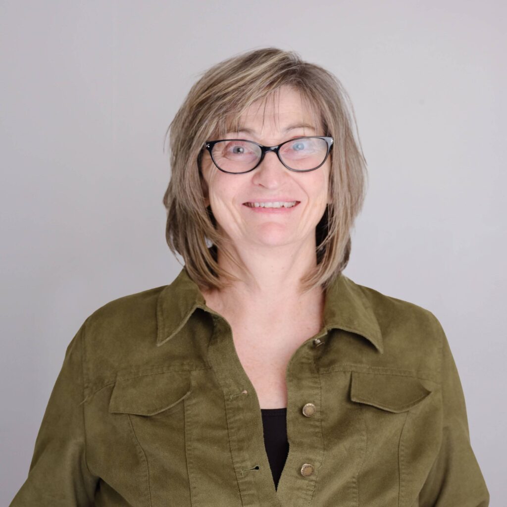 A person with shoulder-length hair and glasses smiles at the camera. They are wearing a green button-up shirt. The background is plain and light-colored.