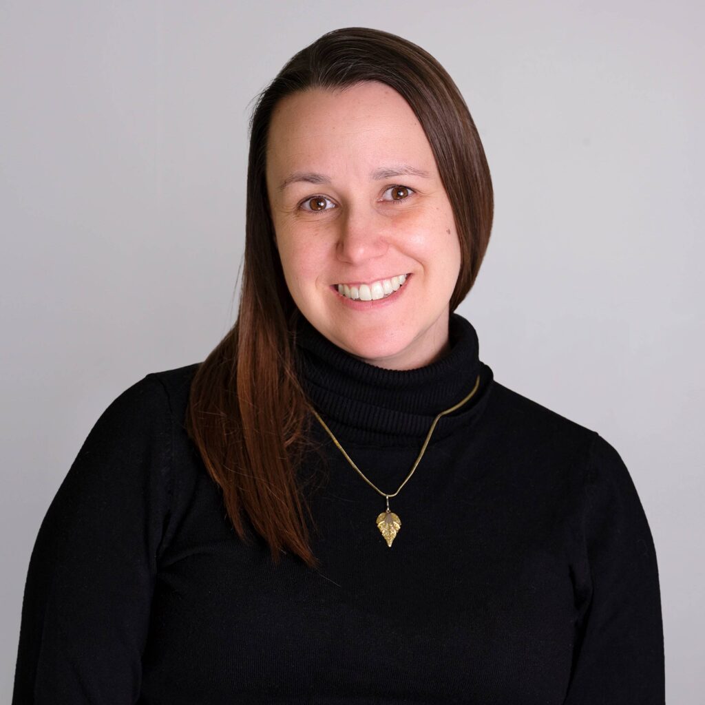 A person with long brown hair is smiling at the camera. They are wearing a black turtleneck and a gold leaf pendant necklace. The background is plain and light-colored.