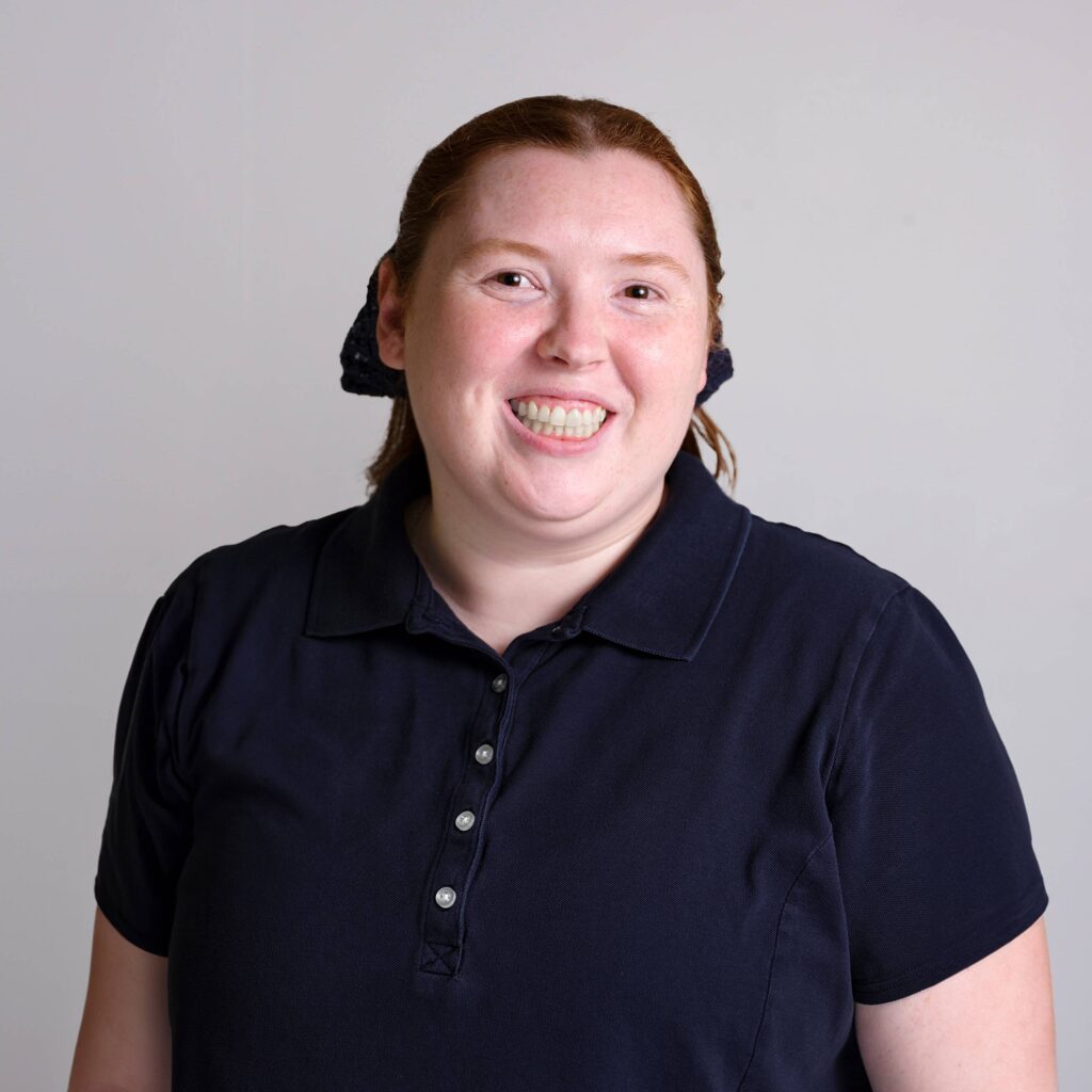 A person with long hair, tied with a scrunchie, smiles warmly. They are wearing a dark navy collared shirt and are standing against a plain light gray backdrop.