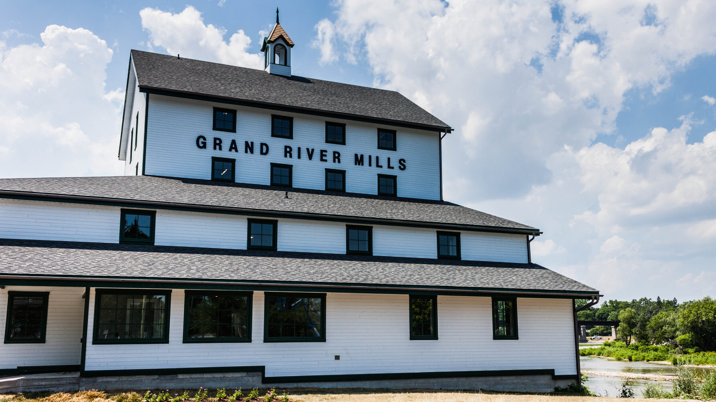 A large, white historic building labeled "Grand River Mills" with several windows and a small tower on the roof, set under a cloudy blue sky. The building is surrounded by greenery and there is a glimpse of a river nearby.