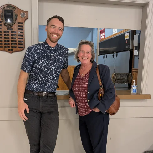 Two people stand smiling in a casual indoor setting. The man on the left wears a patterned shirt and dark pants, while the woman on the right wears a blazer and holds a bag.