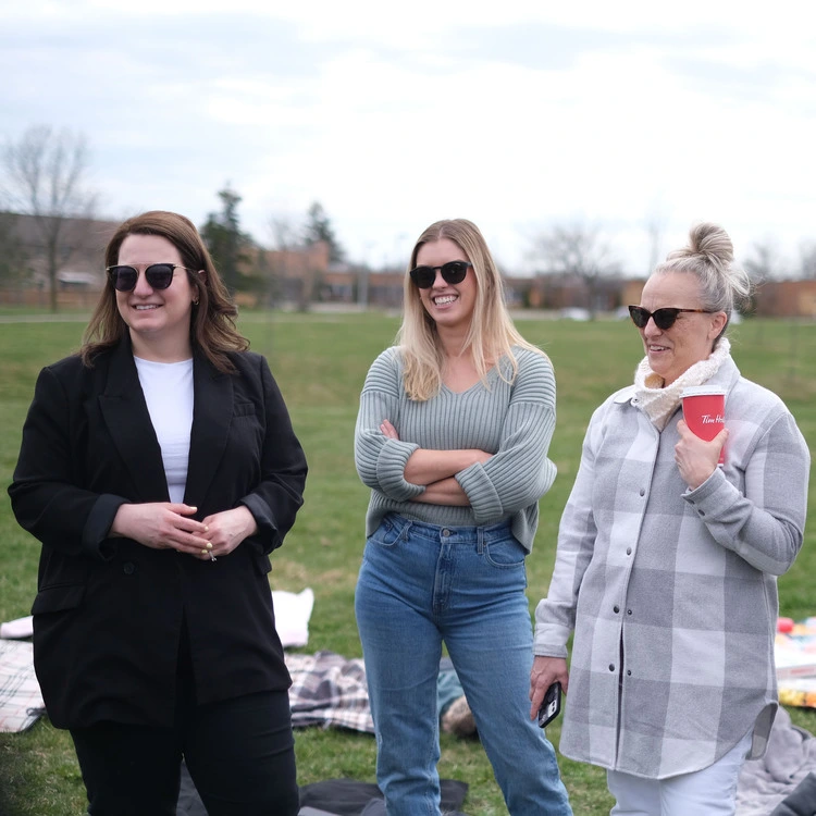 Three women standing outdoors on a grassy field, smiling and wearing sunglasses. One holds a red cup. They are dressed casually, with one in a black jacket, another in a gray sweater, and the third in a checked shirt. It appears to be a cool day.