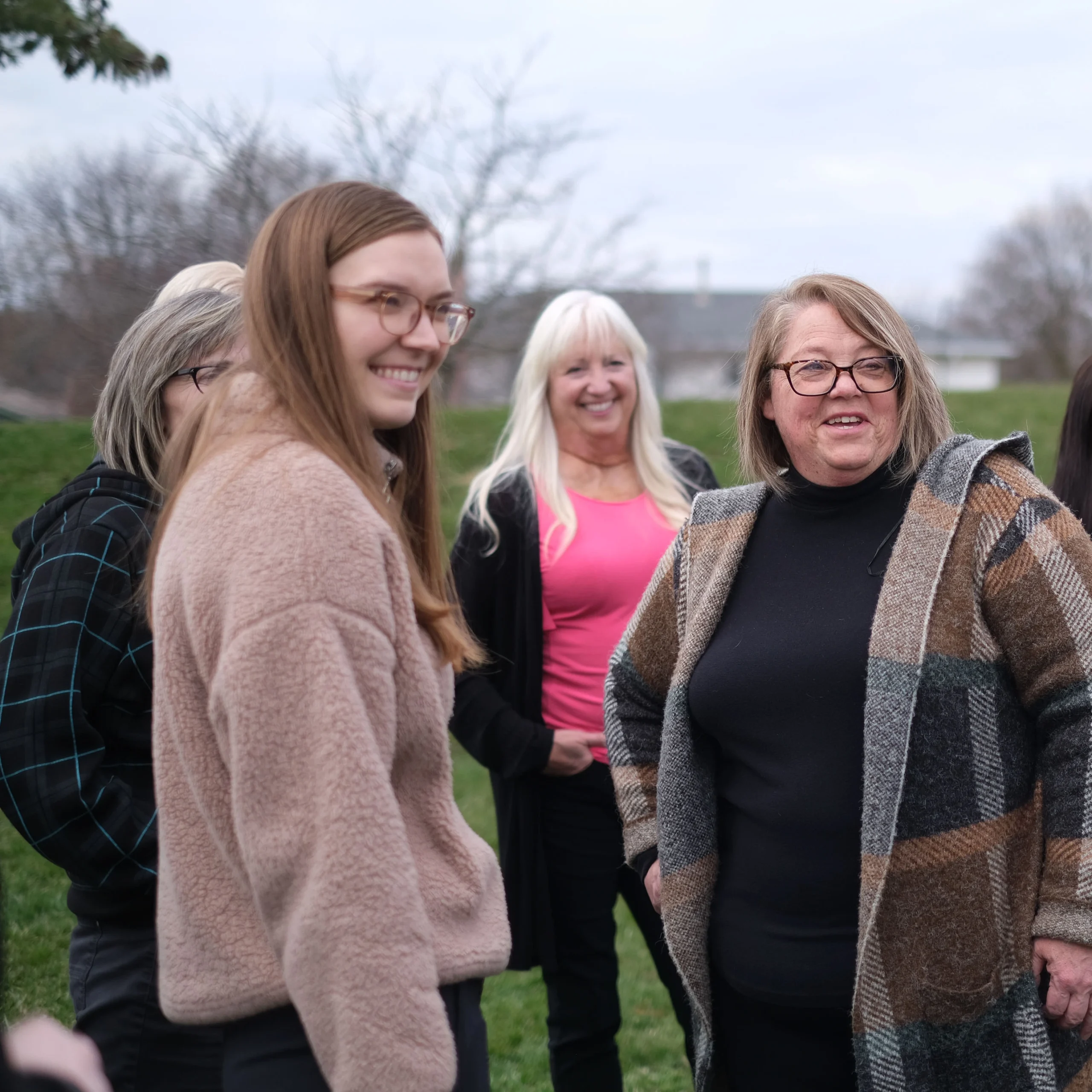 A group of women standing outside, smiling and chatting. They are dressed warmly in jackets and sweaters. The background shows bare trees and a cloudy sky, indicating a cool day.