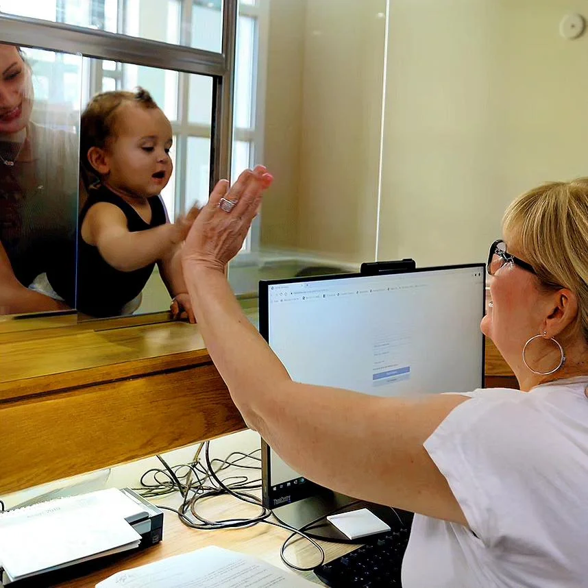 A baby gives a high-five through a glass partition to a smiling woman sitting at a desk with a computer. Another woman holds the baby from behind. The setting appears to be an office or reception area.